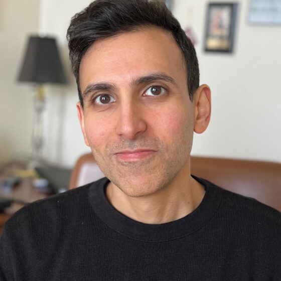 A man with short black hair slightly grinning in a black shirt sitting indoors.