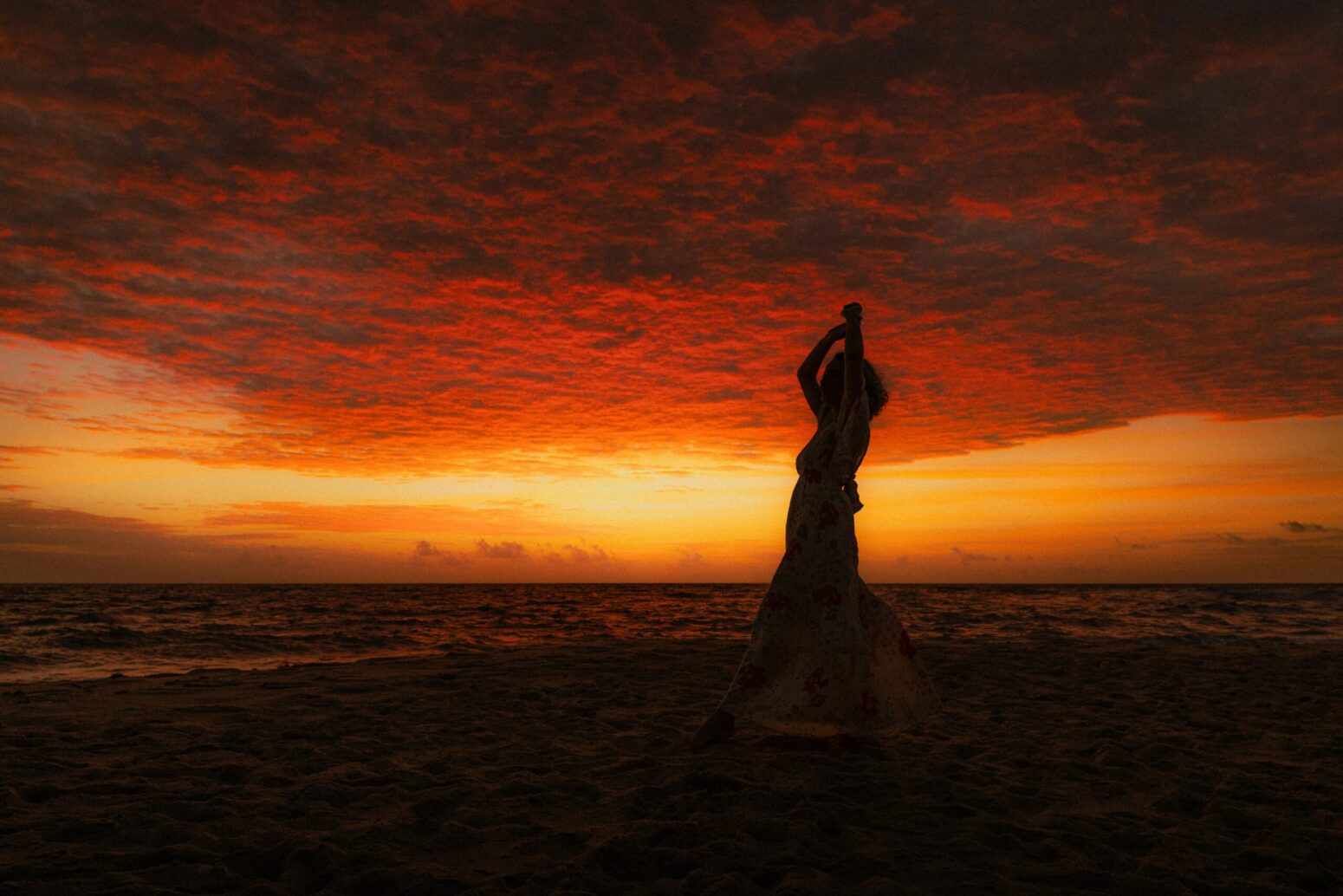 A silhouette of a woman against a orange/red background on a beach with her arms extended above her head