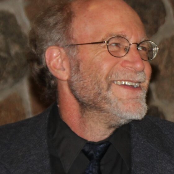 A bald man with graying beard and rectangle classes, smiling and looking to the right