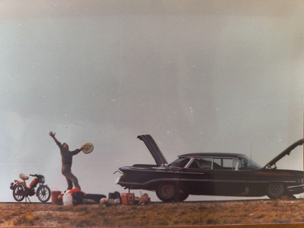 A man dances ecstatically, hat in hand, behind a vintage car with its trunk and hood open. At his feet are things to load into the car. Behind him is a motorbike.