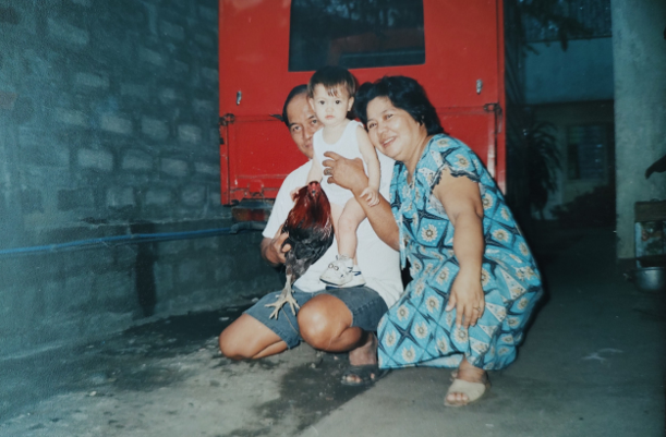 A baby in a white onesie and sneakers and a rooster being held by an older Filipino couple that kneels in a garage-like space.