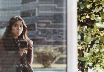 Asa Drake's mother, a young Filipino woman with long hair, photographs her reflection in an outdoor window.