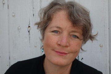 A white woman with short gray hair smiles, her mouth closed, into the camera as she stands in front of a white wall.
