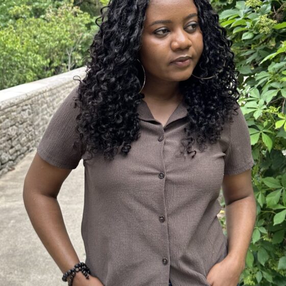 Woman with curly braids standing in the middle of a park. She is wearing a brown shirt, and her hands are tucked into the pockets of her jeans.