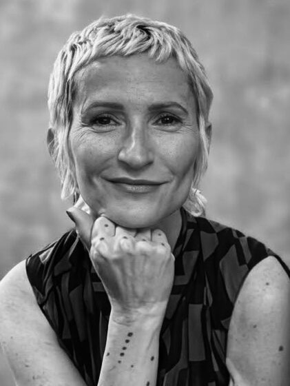 Wearing a sleeveless black shirt, a woman with a short haircut smiles. She poses with her fist below her chin.