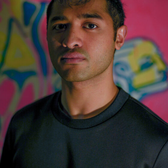 A light-to-middle-brown skin man in a black shirt, standing in front of a graffiti wall. He has a short haircut.