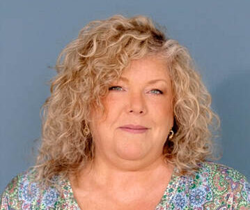 Kathleen Spada, a white woman with wavy blond hair, looks directly into the camera in front of a blue photo backdrop.